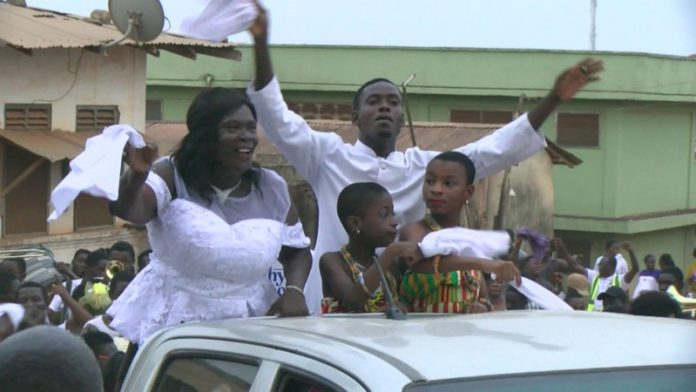 Father Stephen in a plain white cassock, his mother in a white kaba with black stripes and a bright headgear. Father Stephen in a plain white cassock, his mother in a white kaba with black stripes and a bright headgear.