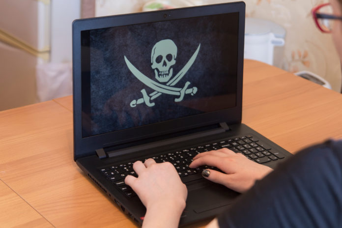 Man or woman hacker or typist, whose face is not visible, holding hands on a laptop keyboard, the screen is a pirate flag Jolly Roger